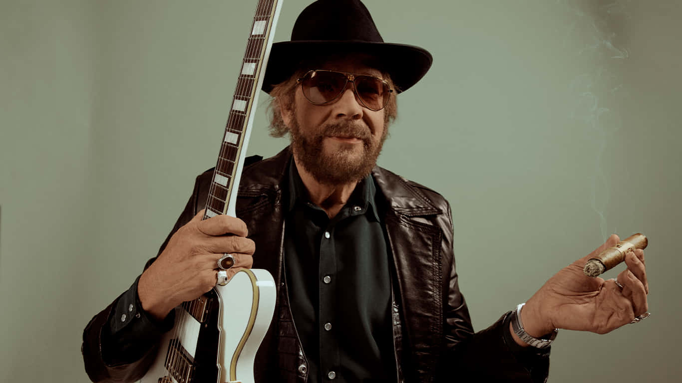 A Man In A Hat And A Jacket Holding A Guitar Wallpaper