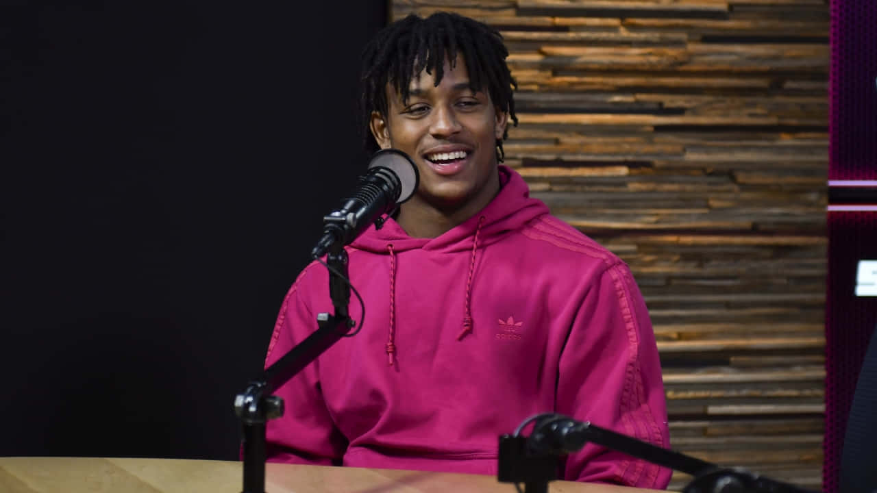 A Man In A Pink Hoodie Is Sitting In Front Of A Microphone Wallpaper