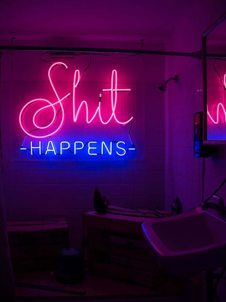 A Neon Sign That Says Shit Happens In A Bathroom Wallpaper