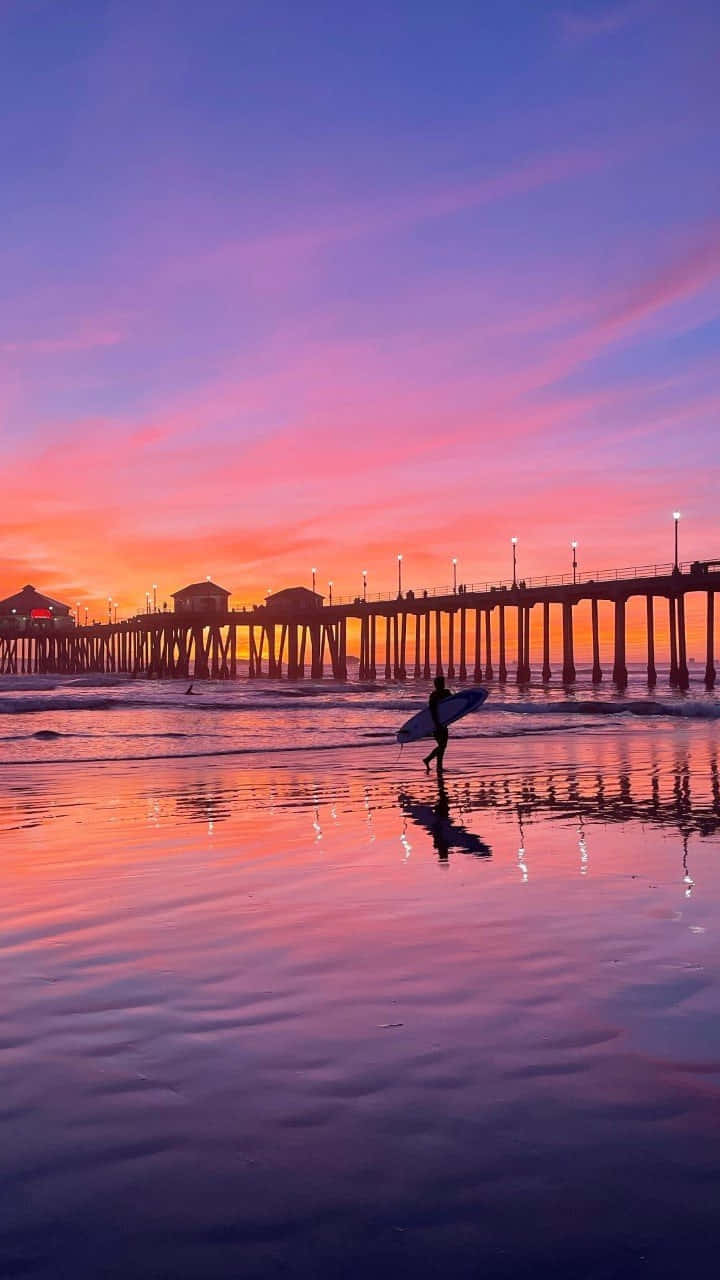 A Surfer Walks Along The Beach At Sunset Wallpaper