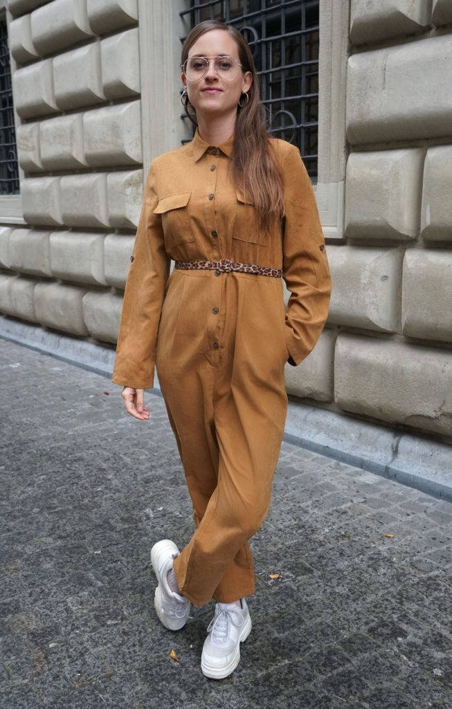 A Woman In A Brown Jumpsuit Standing On A Street Wallpaper