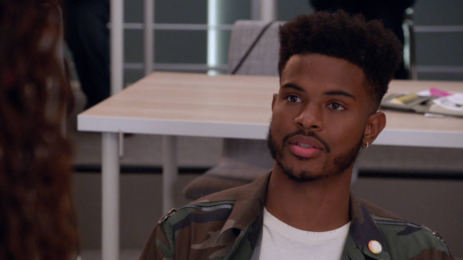 A Young Man In A Camouflage Jacket Is Sitting In An Office Wallpaper