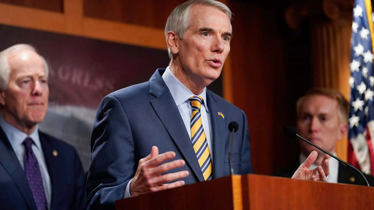 Rob Portman Speaking Two Men Behind Him Wallpaper