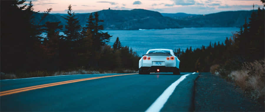 A White Car Driving Down A Road Near A Lake Wallpaper