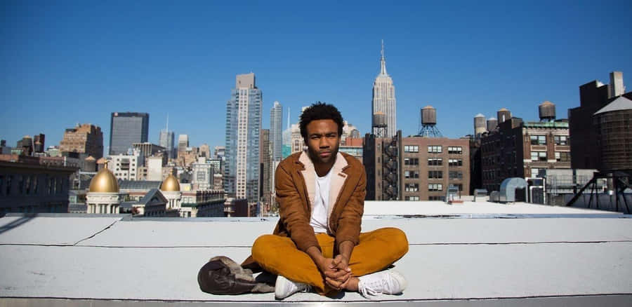 Donald Glover Sittingon Rooftop With City Backdrop Wallpaper