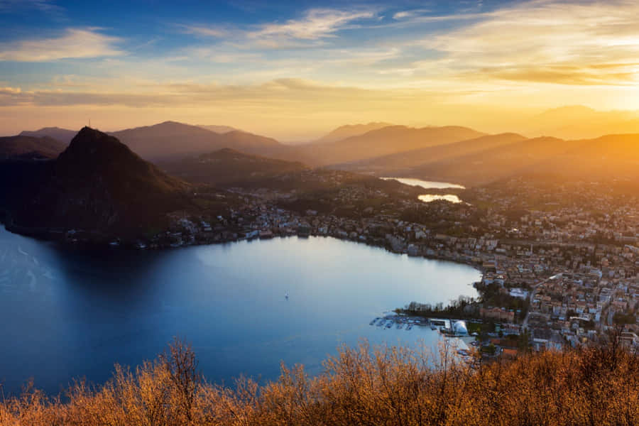 Stunning View Of Lugano Lake Surrounded By Majestic Mountains Wallpaper