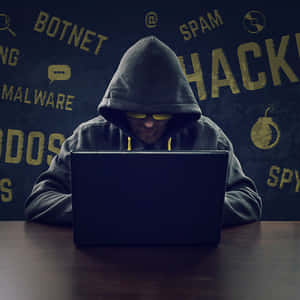 A Man In A Hoodie Is Sitting At A Table With A Laptop Wallpaper