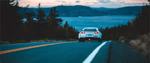 A White Car Driving Down A Road Near A Lake Wallpaper