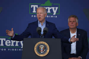 Joe Biden Delivering A Speech With Terry Mcauliffe Behind Him Wallpaper