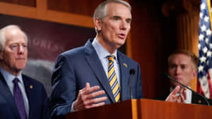 Rob Portman Speaking Two Men Behind Him Wallpaper