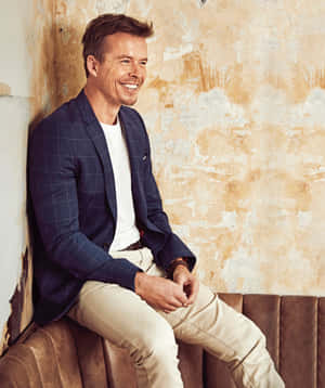 Smiling Manin Blue Blazer Sittingon Couch Wallpaper